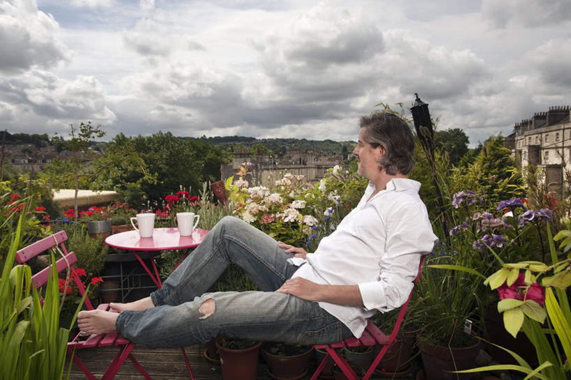 西方人,一个人,家,坐,笑,微笑,露齿笑,横图,室外,白天,侧面,草地,草坪,山,山脉,植物,桌面,桌子,杯子,椅子,欧洲,欧洲人,仅一个男性,仅一个人,仅一个中年男性,阴影,服装,盛开,光线,木制,影子,陶瓷,房屋,墙,屋子,山峰,云,云朵,陶瓷制品,山峦,花蕾,云彩,注视,休闲装,楼房,墙壁,墙面,住宅,草,花,花瓣,花朵,鲜花,树,树木,两个,衣服,蓝色,绿色,白色,白云,蓝天,天空,阳光,自然,马克杯,木制品,水杯,群山,天,花苞,花蕊,观察,看,休闲,休闲服,搭,服饰,生长,晴朗,成长,观看,察看,搭着,关注,男人,男性,中年男性,白种人,晴空,坐着,彩图,全身,房子